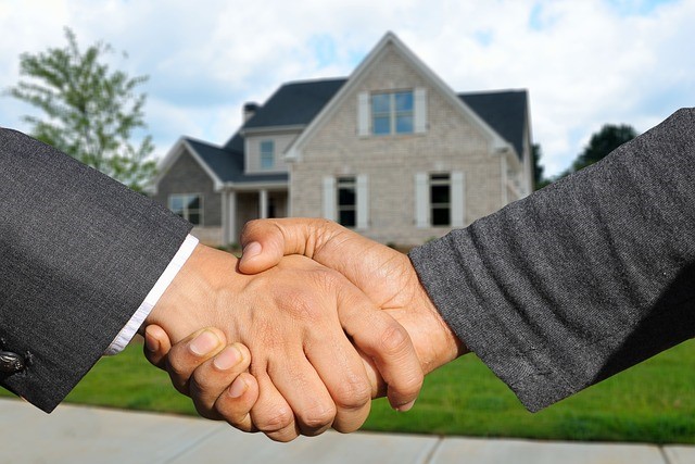 Two people shaking hands in front of a house, symbolizing a real estate agreement or deal.

